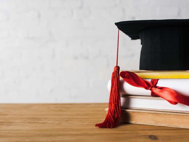 Diploma with beautiful bow, graduation cap with red tassel on top of books on table on white